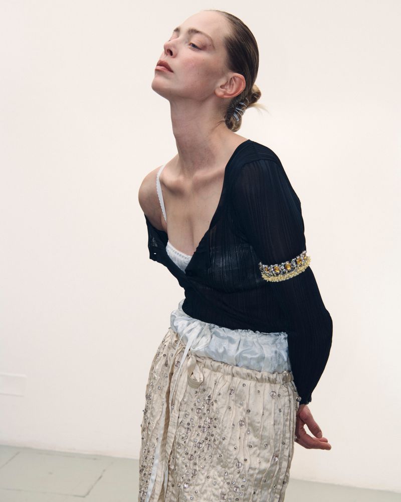 Model with pulled back hair wearing a black sheer top and embellished trousers against a white seamless background