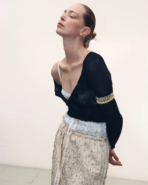 Model with pulled back hair wearing a black sheer top and embellished trousers against a white seamless background