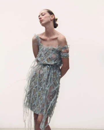 Model wearing a sheer blue embellished dress with ribbons and beaded detailing against a plain white background