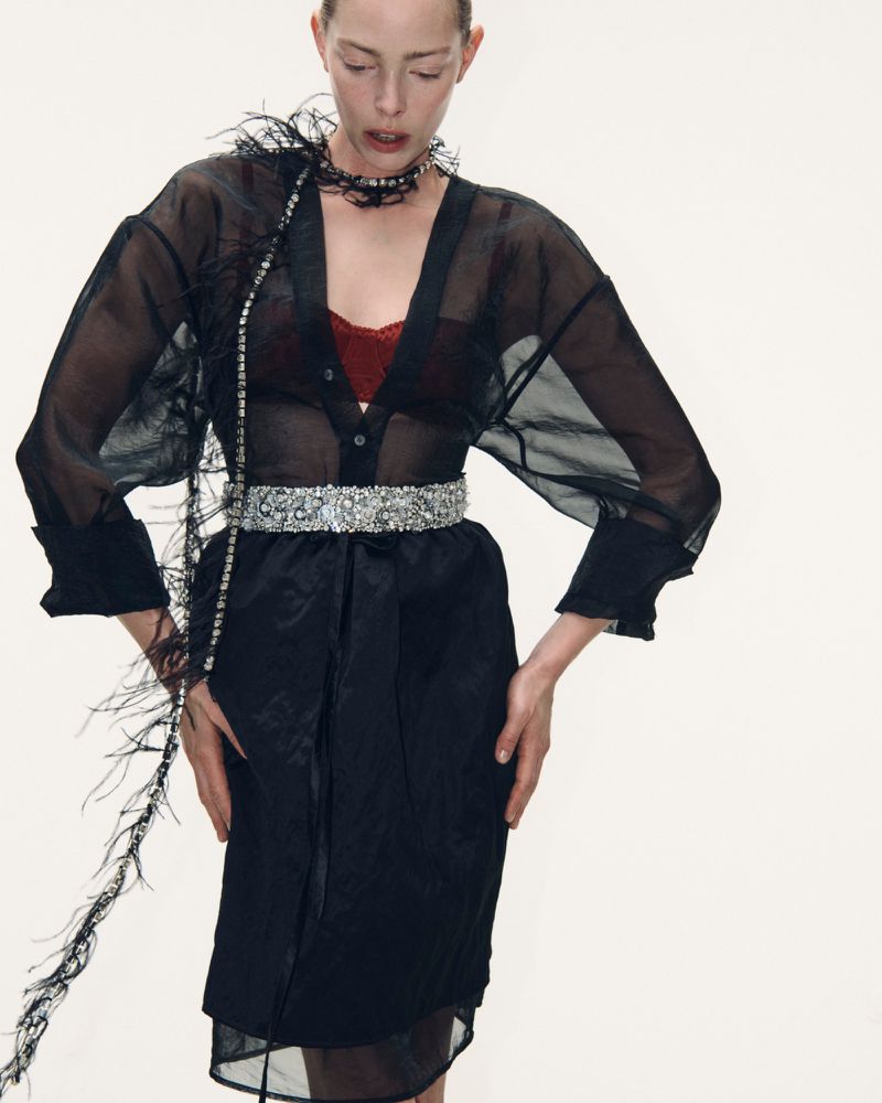 Model Tanya Diagileva wearing a sheer black button-down shirt and embellished belt against a white seamless background