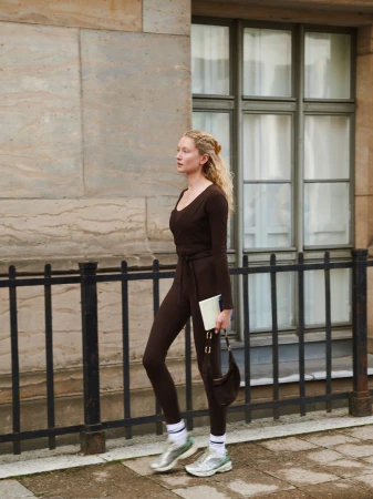 Model in brown activewear holding a notebook and bag while walking past a building with windows.