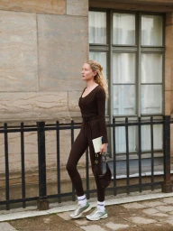 Model in brown activewear holding a notebook and bag while walking past a building with windows.
