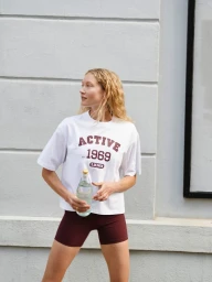 Model wearing a white t-shirt and maroon shorts holding a water bottle outdoors against a building exterior.