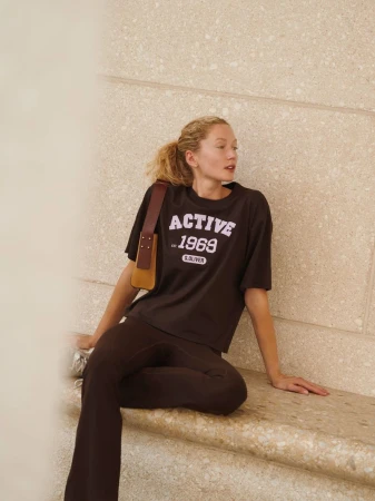 Model wearing dark t-shirt with text graphic and brown trousers sitting against a stone wall
