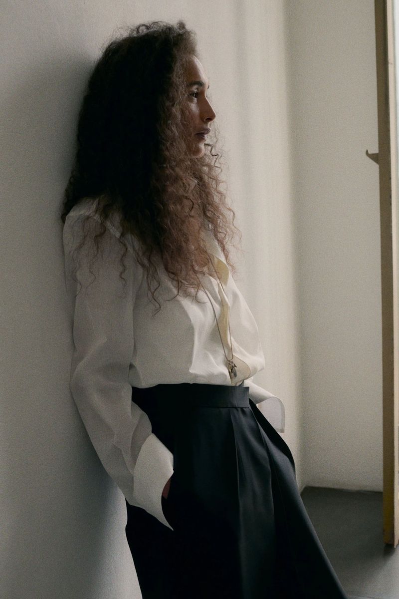Model with long curly hair leaning against a plain wall wearing a white button-down shirt and black trousers.