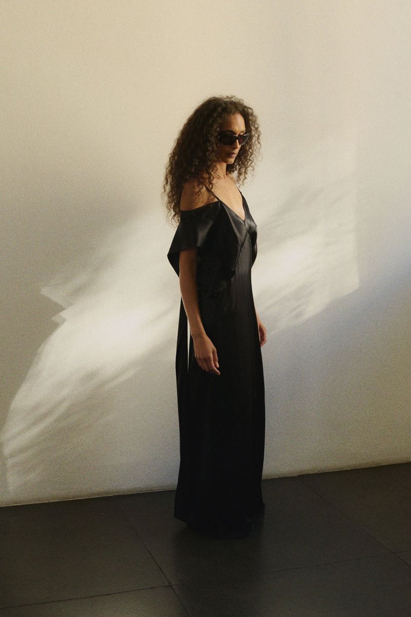 Model with curly hair wearing dark sunglasses and a long black dress standing against a textured wall with light cast across it.