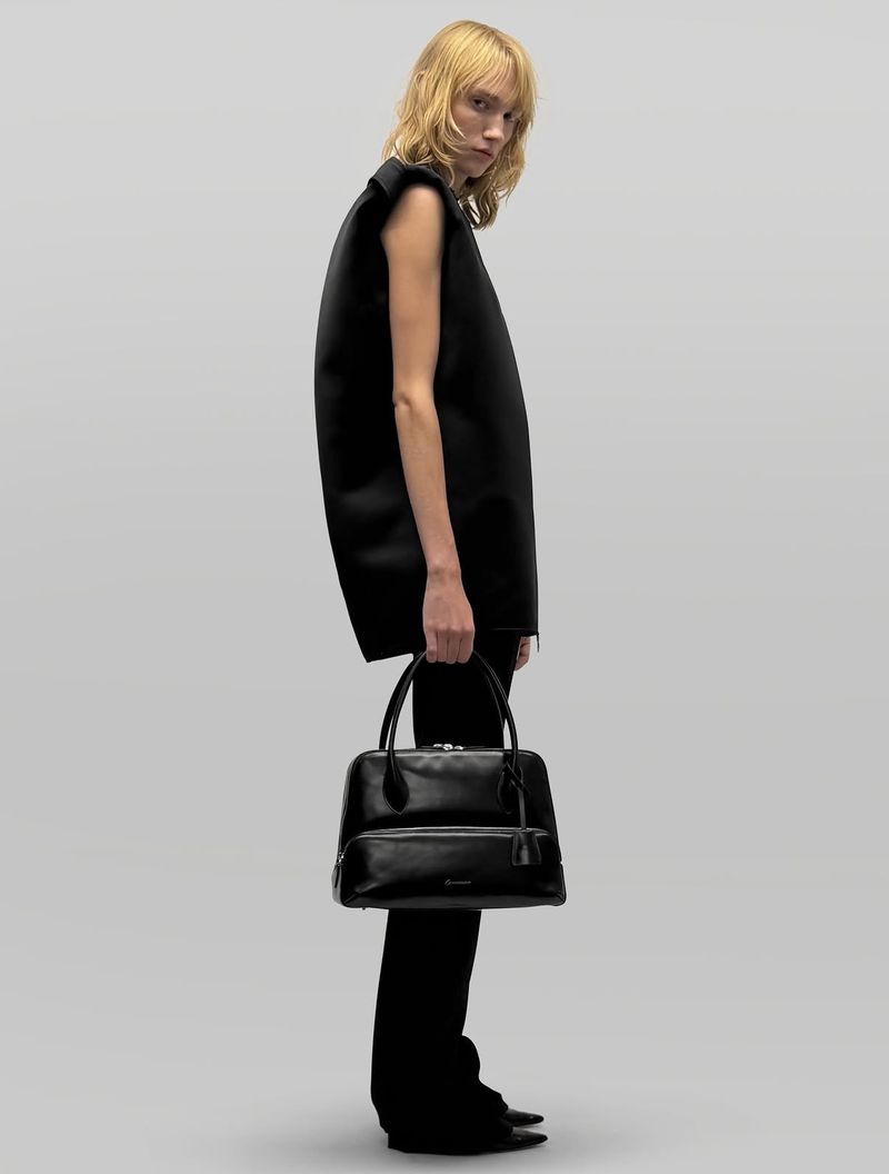 Model in black top and trousers holding a leather handbag against a light grey studio background