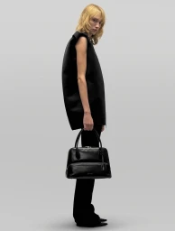Model in black top and trousers holding a leather handbag against a light grey studio background
