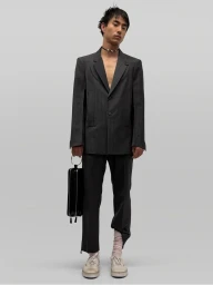 Model wearing a dark pinstripe suit holding a black structured handbag against a seamless grey backdrop.