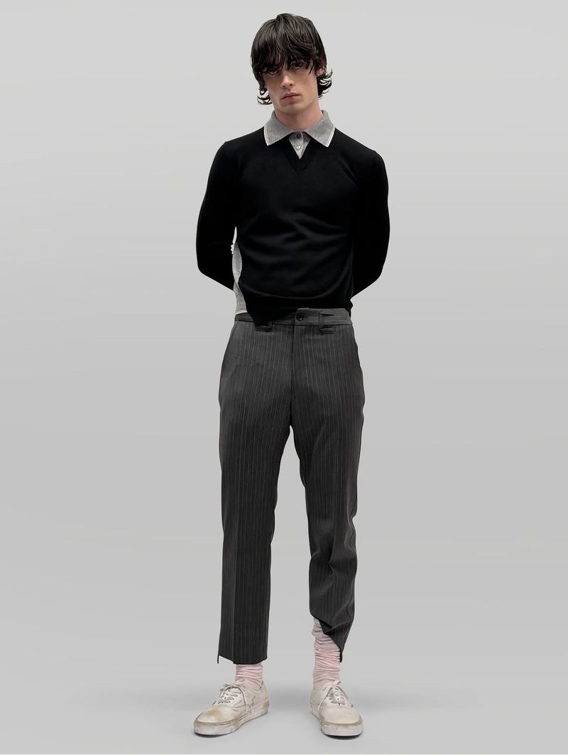 Full-length portrait of a male model wearing a black sweater with a grey collar, striped trousers, and white sneakers against a neutral grey backdrop.
