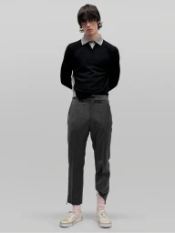 Full-length portrait of a male model wearing a black sweater with a grey collar, striped trousers, and white sneakers against a neutral grey backdrop.