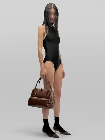 Model in a black bodysuit holding a brown leather handbag against a neutral grey studio seamless backdrop.