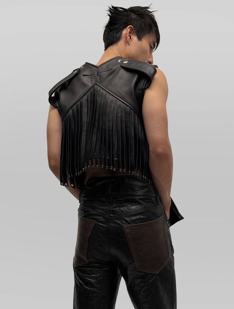 Man wearing a black leather fringe vest and matching trousers against a neutral grey studio background