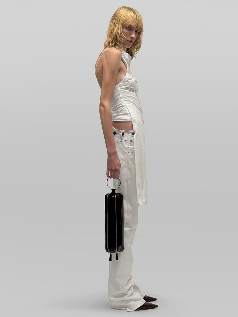 Model in white top and trousers holding a black cylindrical handbag against a grey seamless background