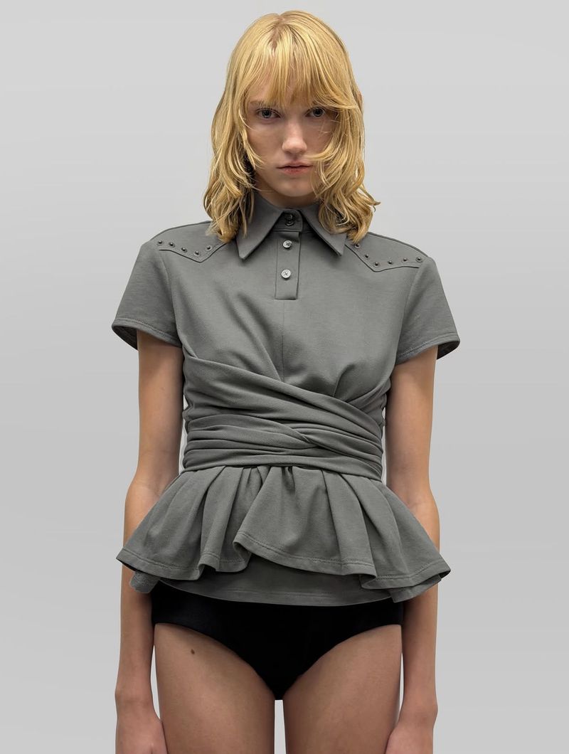 Model with blonde hair wearing a grey polo shirt with a ruffled peplum waist against a plain light grey background.
