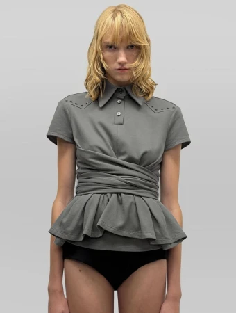 Model with blonde hair wearing a grey polo shirt with a ruffled peplum waist against a plain light grey background.