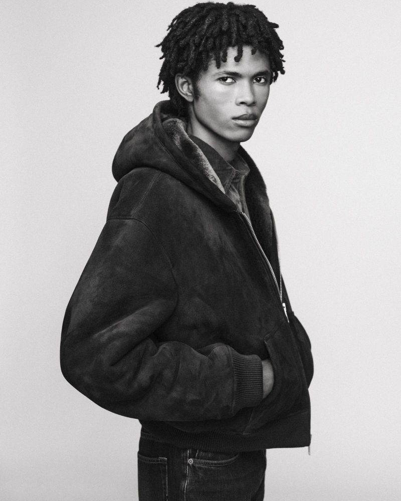 Monochrome portrait of a young male model wearing a dark hooded jacket against a plain background for AllSaints F/W 26 lookbook.