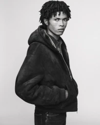 Monochrome portrait of a young male model wearing a dark hooded jacket against a plain background for AllSaints F/W 26 lookbook.