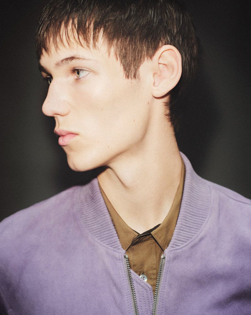 Profile portrait of a young male model wearing a light purple bomber jacket over an olive collared shirt against a dark background