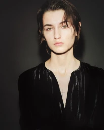 Close-up portrait of a model with short dark hair wearing a black top against a dark seamless backdrop.