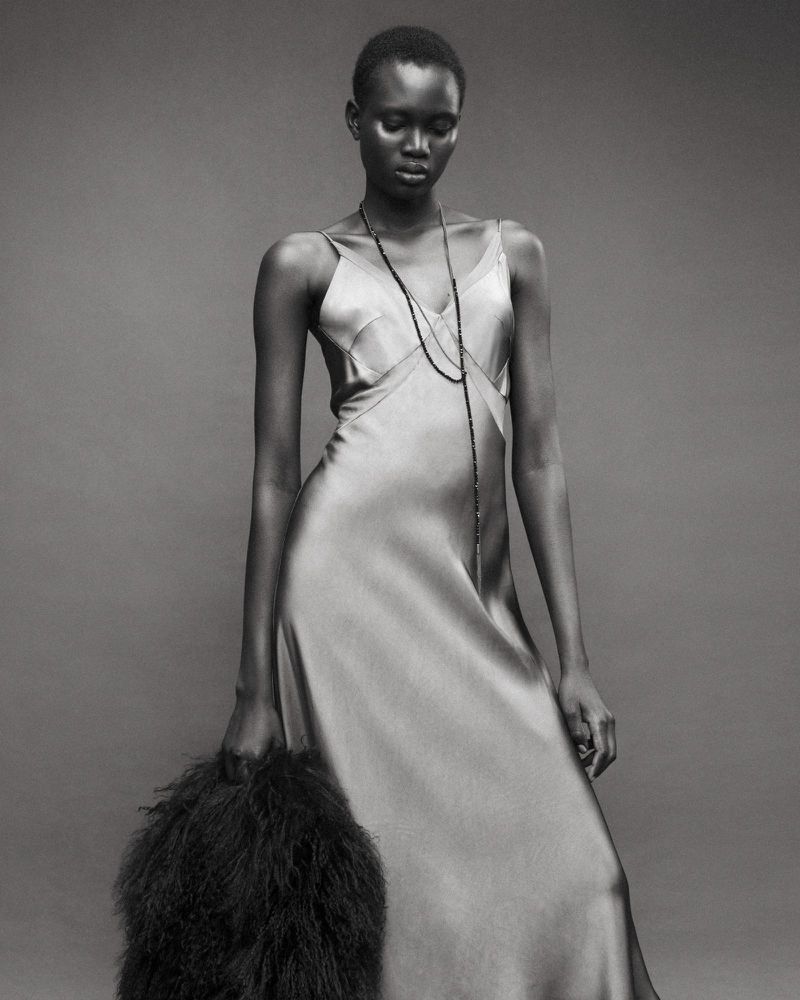 Model wearing a glossy silk slip dress and long necklace holding a furry bag against a plain studio grey backdrop.