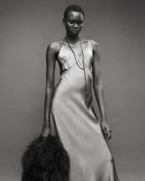 Model wearing a glossy silk slip dress and long necklace holding a furry bag against a plain studio grey backdrop.