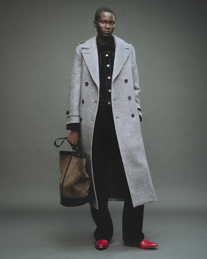 Model wearing a grey double-breasted long coat holding a bucket bag against a grey seamless background