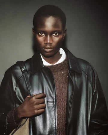 Portrait of a model wearing a dark leather jacket and brown knit sweater against a plain dark grey studio background.