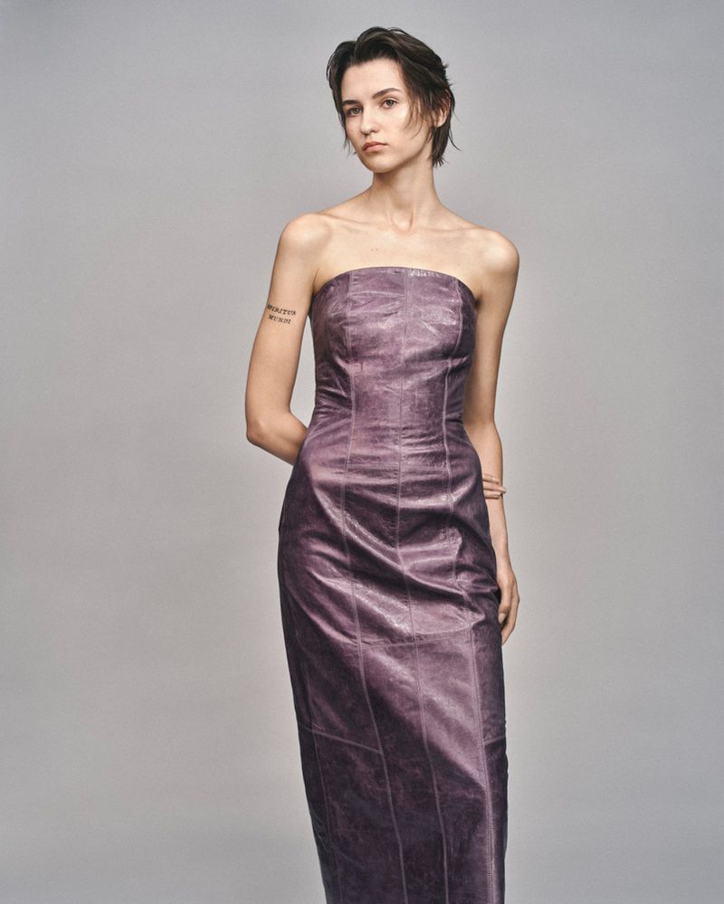 Model with short dark hair wearing a strapless purple midi dress against a plain grey studio backdrop