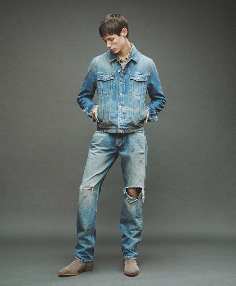 Male model in a distressed denim jacket and ripped jeans against a grey backdrop for AllSaints Fall Winter 26