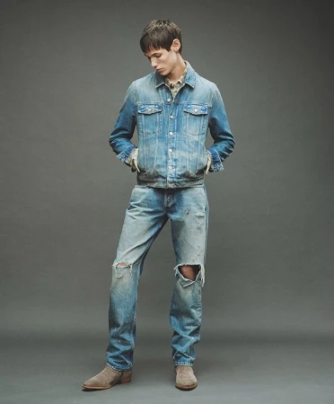 Male model in a distressed denim jacket and ripped jeans against a grey backdrop for AllSaints Fall Winter 26