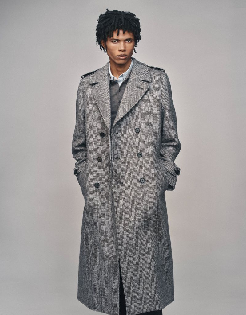 Model wearing a grey wool double-breasted trench coat with hands in pockets against a plain studio background.