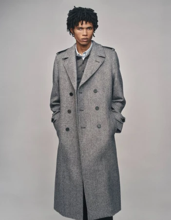 Model wearing a grey wool double-breasted trench coat with hands in pockets against a plain studio background.