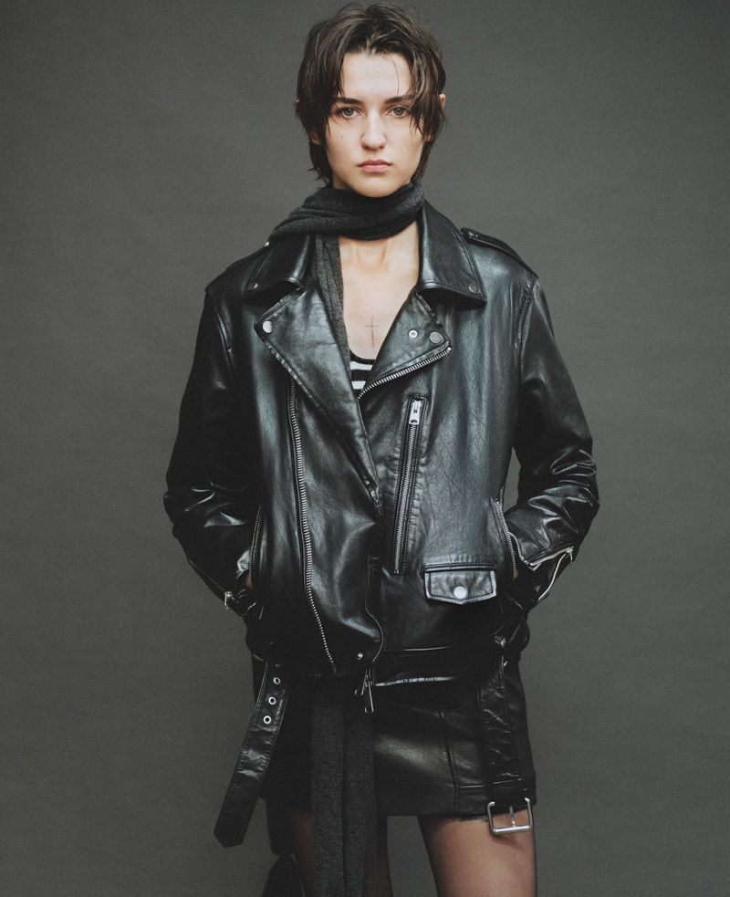 Model in black leather jacket and matching skirt standing against a dark grey seamless studio background.