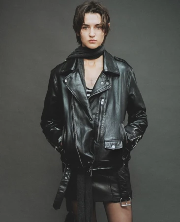 Model in black leather jacket and matching skirt standing against a dark grey seamless studio background.