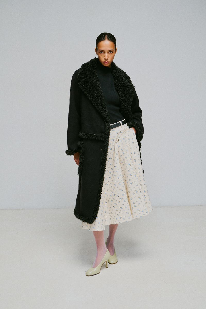 Model wearing a black shearling-trimmed coat and patterned midi skirt against a plain light grey studio backdrop.