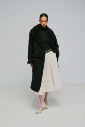 Model wearing a black shearling-trimmed coat and patterned midi skirt against a plain light grey studio backdrop.