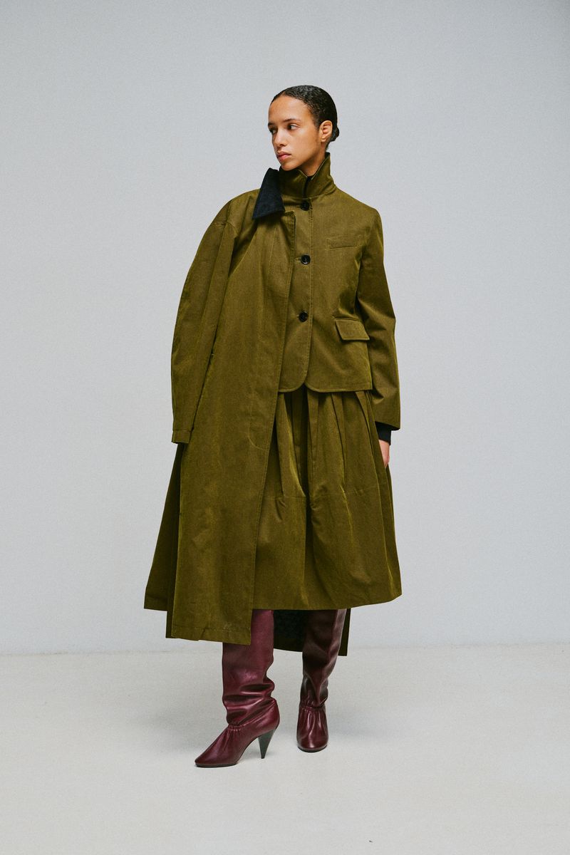 Model wearing an olive green coat and skirt set with burgundy boots against a plain grey studio backdrop