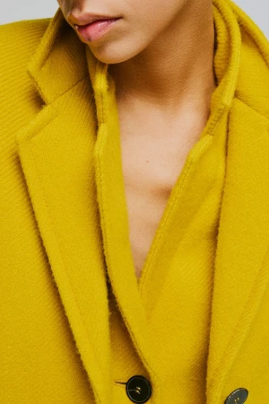 Close-up portrait of a model wearing a textured mustard yellow wool coat with dark buttons against a neutral grey background.