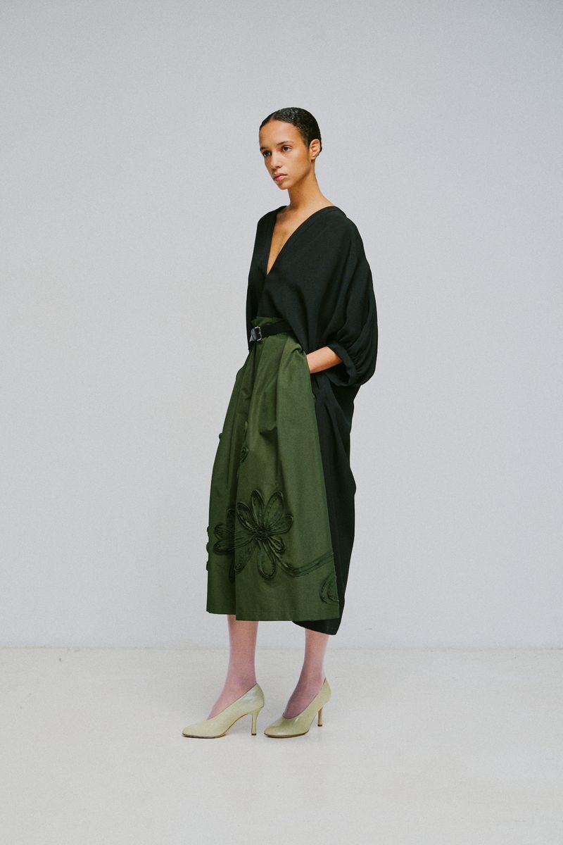 Model standing against a white seamless background wearing a black v-neck top and an olive green pleated skirt.