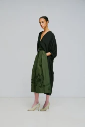 Model standing against a white seamless background wearing a black v-neck top and an olive green pleated skirt.