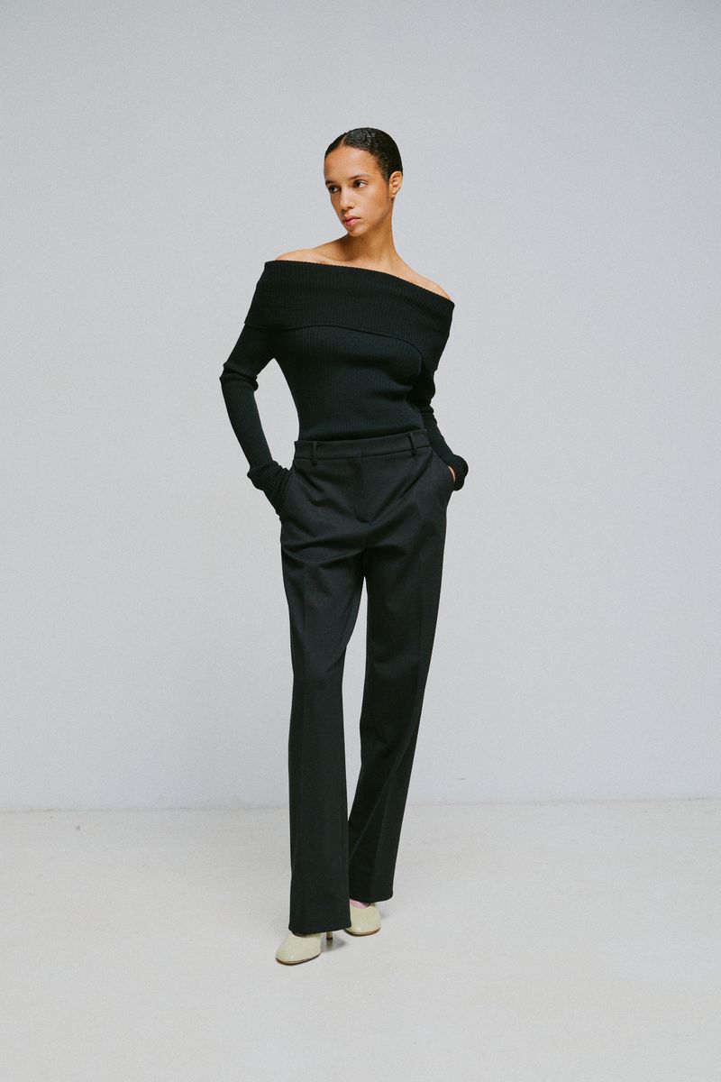 Model wearing an off-the-shoulder black top and dark trousers against a plain grey studio backdrop