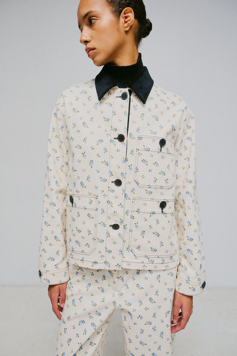Model wearing a cream floral print jacket and matching trousers with a dark collar against a plain studio background.