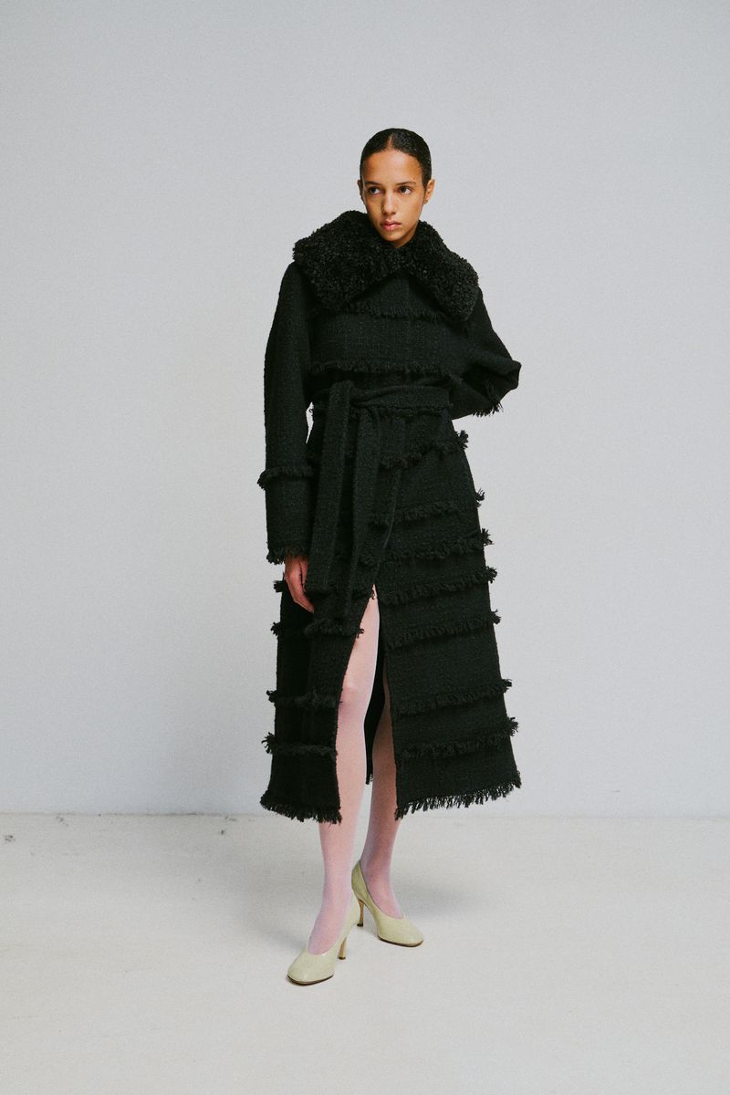 Model wearing a long black fringed coat with a furry collar and light pink tights against a grey studio wall background
