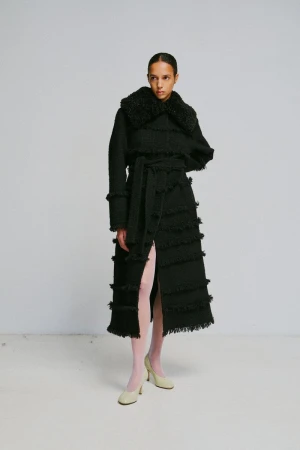 Model wearing a long black fringed coat with a furry collar and light pink tights against a grey studio wall background