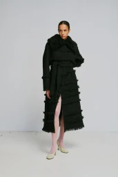 Model wearing a long black fringed coat with a furry collar and light pink tights against a grey studio wall background