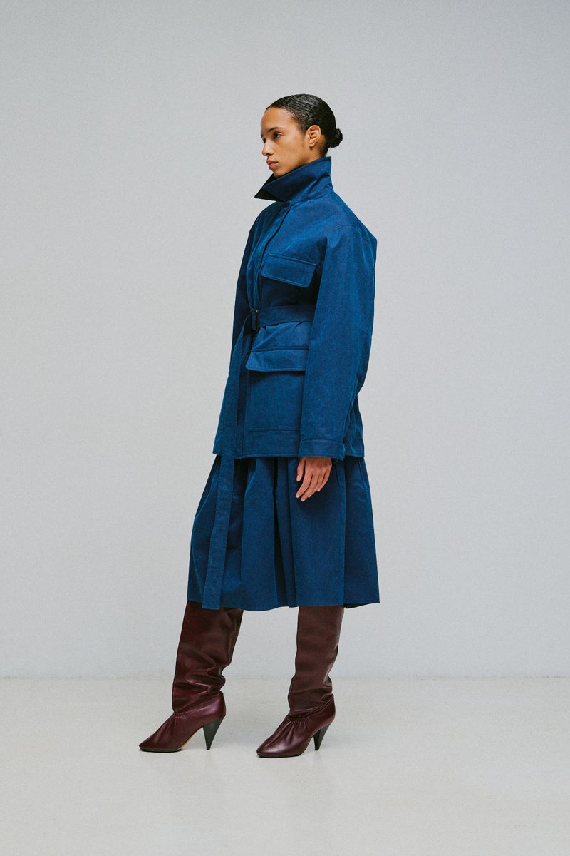Model wearing a blue belted jacket and skirt with burgundy knee-high boots against a plain grey background