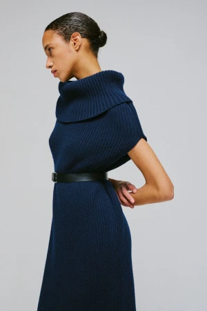 Model wearing a blue ribbed turtleneck dress with a black belt against a grey studio backdrop