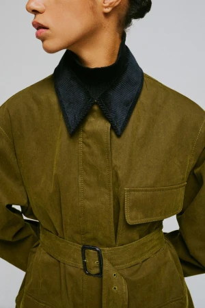 Close-up of a model wearing an olive green belted jacket with a dark corduroy collar against a plain grey background.