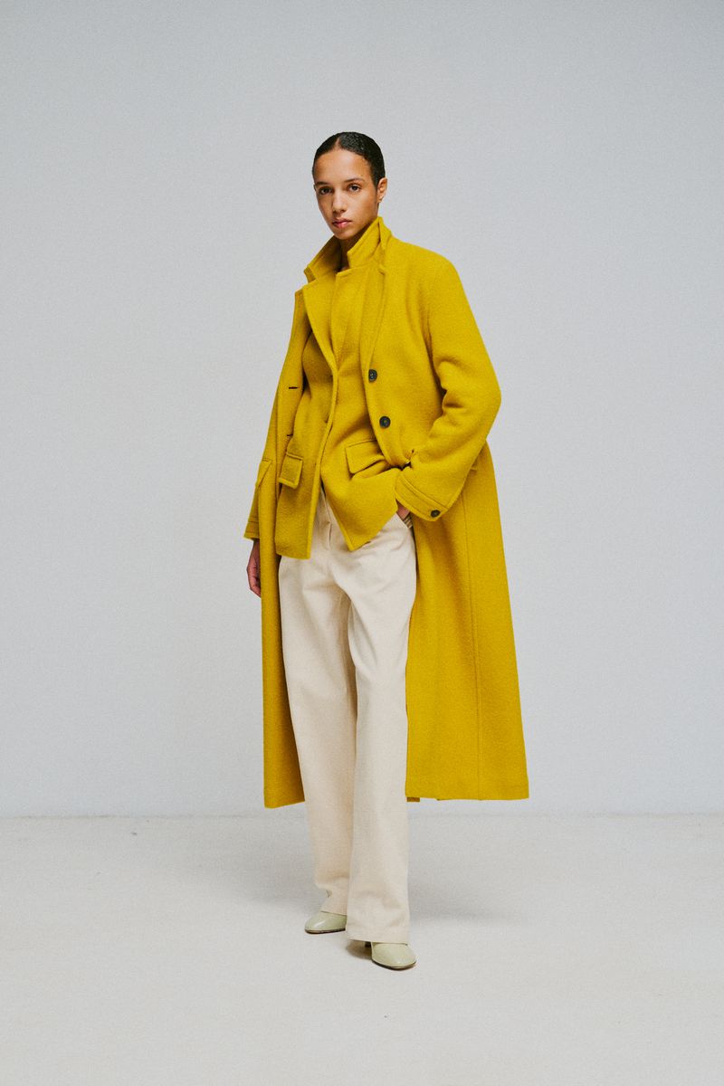 Model wearing a vibrant yellow coat and cream trousers against a neutral grey studio background